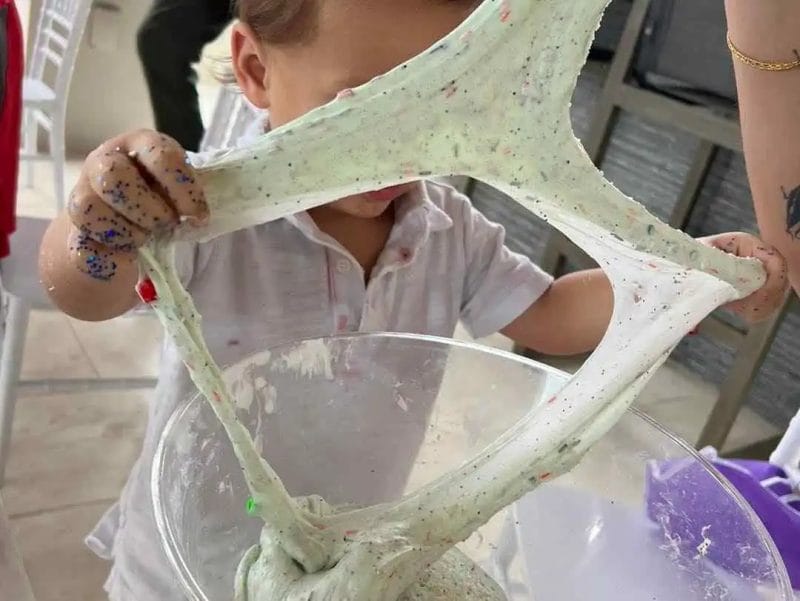 A child stretches homemade green slime with colorful speckles between their hands at a kids party, over a bowl on a table.