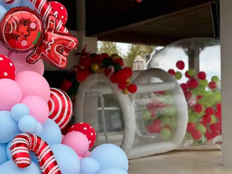 Colorful entrance decorated for a Wonderland event with multi-colored balloons and festive ornaments, including candy cane decorations, with a blurry view of an indoor space through a clear, balloon-decorated doorway.