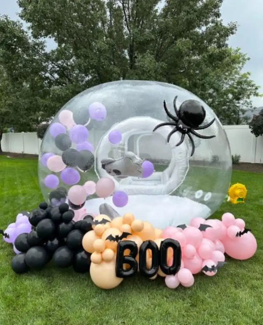 A Halloween-themed outdoor setup with a large transparent bubble filled with purple and white balloons, surrounded by black and pink balloon clusters at a "Balloon House" event.