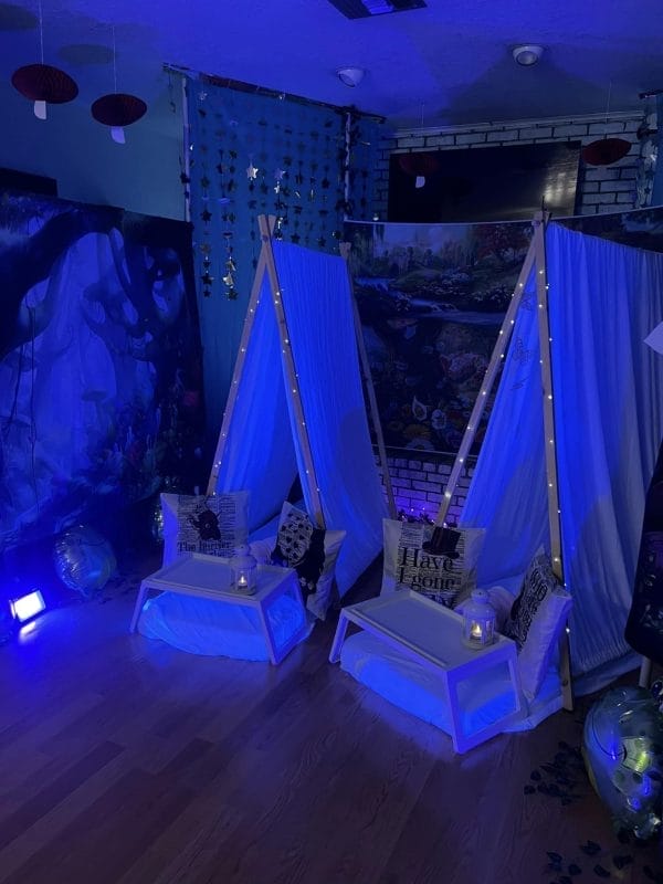 Indoor camping setup with two teepees, fairy lights, and Alice in Wonderland-themed decorations under a blue light.