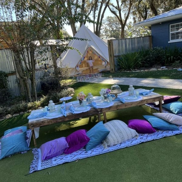 An outdoor children's sleepover party setup featuring a decorative tent with a low table surrounded by colorful cushions and blankets on a lawn in Florida.