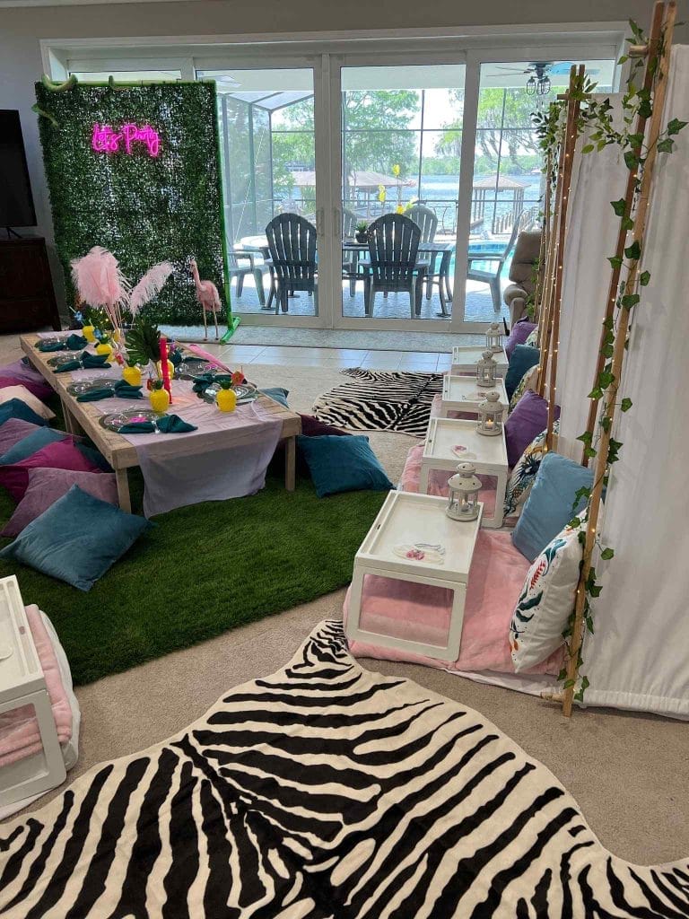 A brightly decorated indoor party area with a tropical theme, including a grass-like carpet, an ultimate sleepover upgrade with a table set with cushions for seating, and flamingo accents.