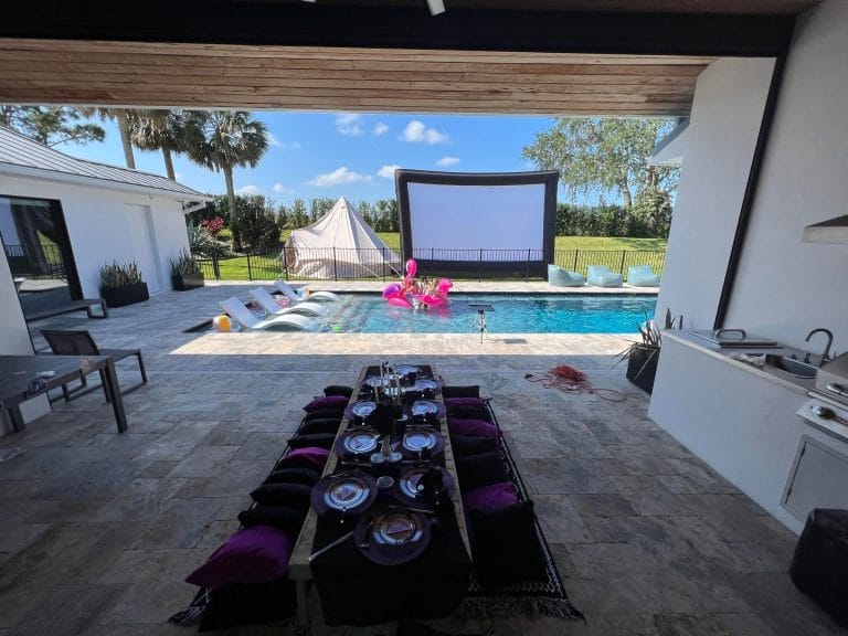 Outdoor dining area with a set table, leading to a pool with inflatable flamingos and a screen for an Ultimate Night movie, surrounded by palm trees.