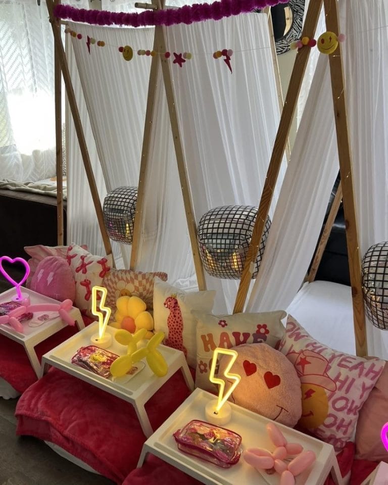 Indoor glamping setup for a slumber party with two beds, a canopy, festive lights, and decorative pillows accompanied by trays of snacks and cushions on the floor.