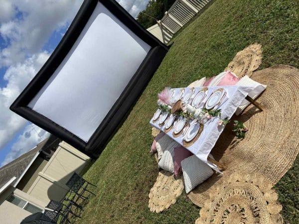 Outdoor movie setup with a large screen and a decorated table with plates and cushions on the grass for the ultimate night.