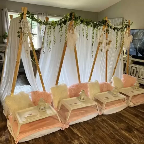Indoor party setup featuring three wooden teepees draped with white fabric and adorned with greenery, positioned above cushioned seating and wooden serving trays for a Barbie Themed Party.