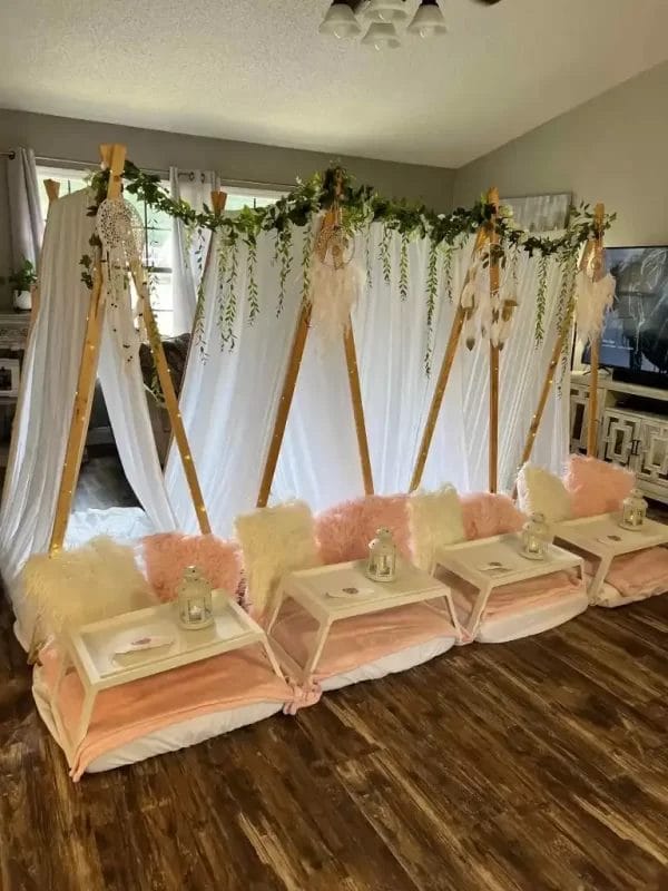 Indoor party setup featuring three wooden teepees draped with white fabric and adorned with greenery, positioned above cushioned seating and wooden serving trays for a Barbie Themed Party.