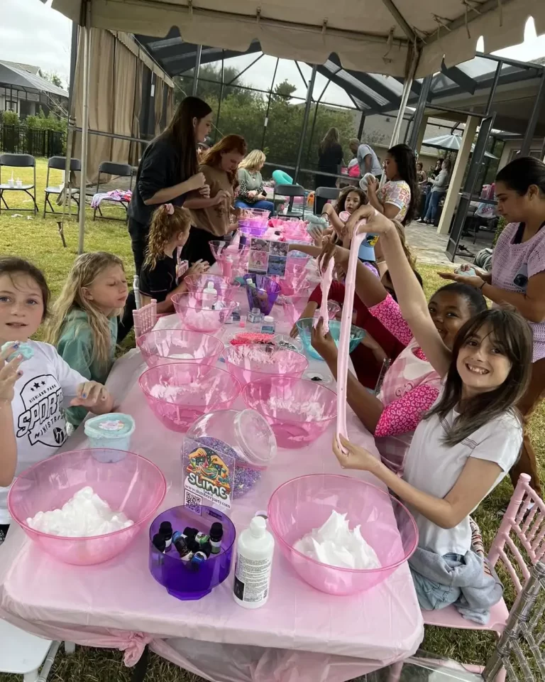 Children and adults gather around tables with pink tablecloths, engaged in making slime at a Barbie Themed Party.