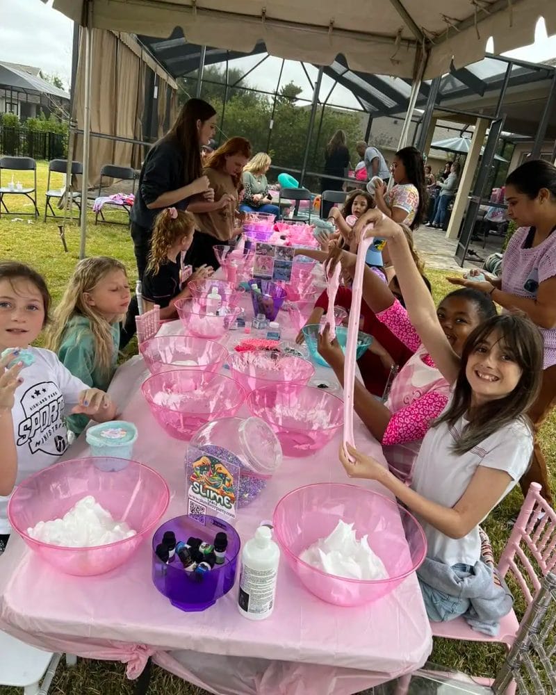 Children and adults gather around tables with pink tablecloths, engaged in making slime at a Barbie Themed Party.