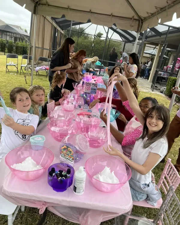 Children enjoying a slime-making activity at an outdoor Barbie Themed Party with pink decorations and supplies.