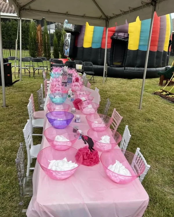 Long table set with pink bowls of white cotton candy, chairs aligned on one side, on a grassy area with Barbie Themed Party decorations and a colorful inflatable castle in the background.