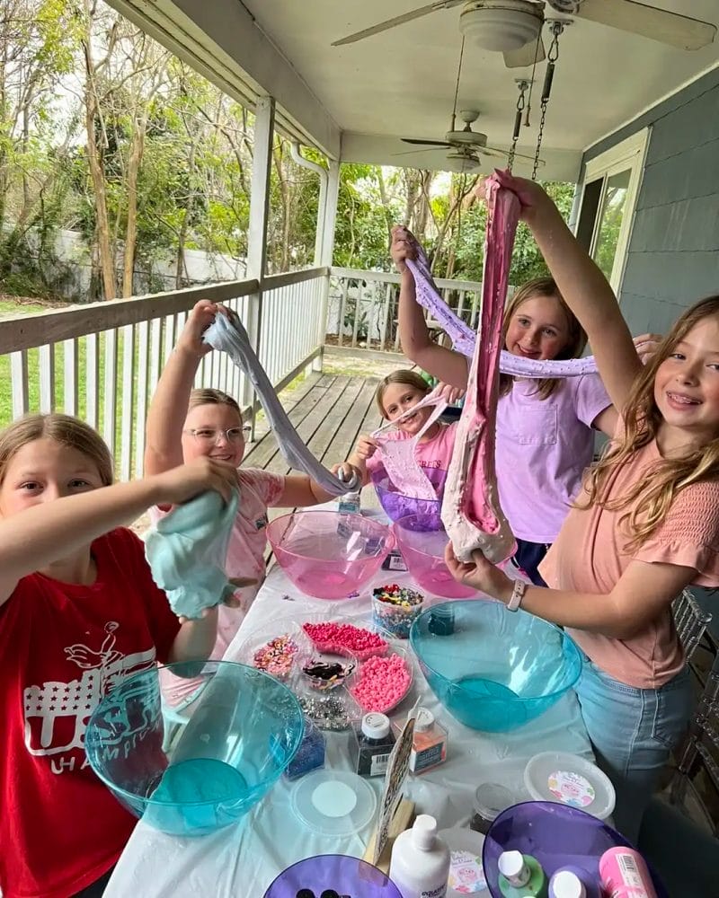 Four children making slime at a Barbie Themed Party on a porch, surrounded by bowls and ingredients, displaying their colorful creations.
