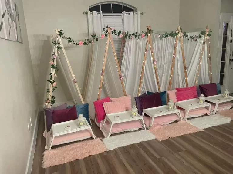 Indoor Barbie Themed Party setup with wooden teepees adorned with fairy lights and floral decorations, lined with colorful cushions and cozy rugs on a hardwood floor.