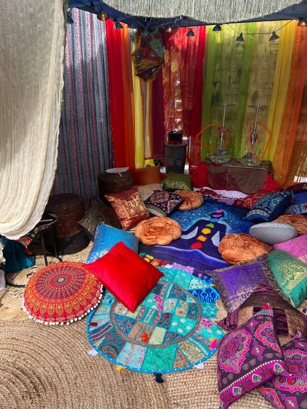 An elaborately decorated tent interior with colorful cushions, rugs, and hanging textiles, creating a vibrant Wonderland-inspired bohemian setting.