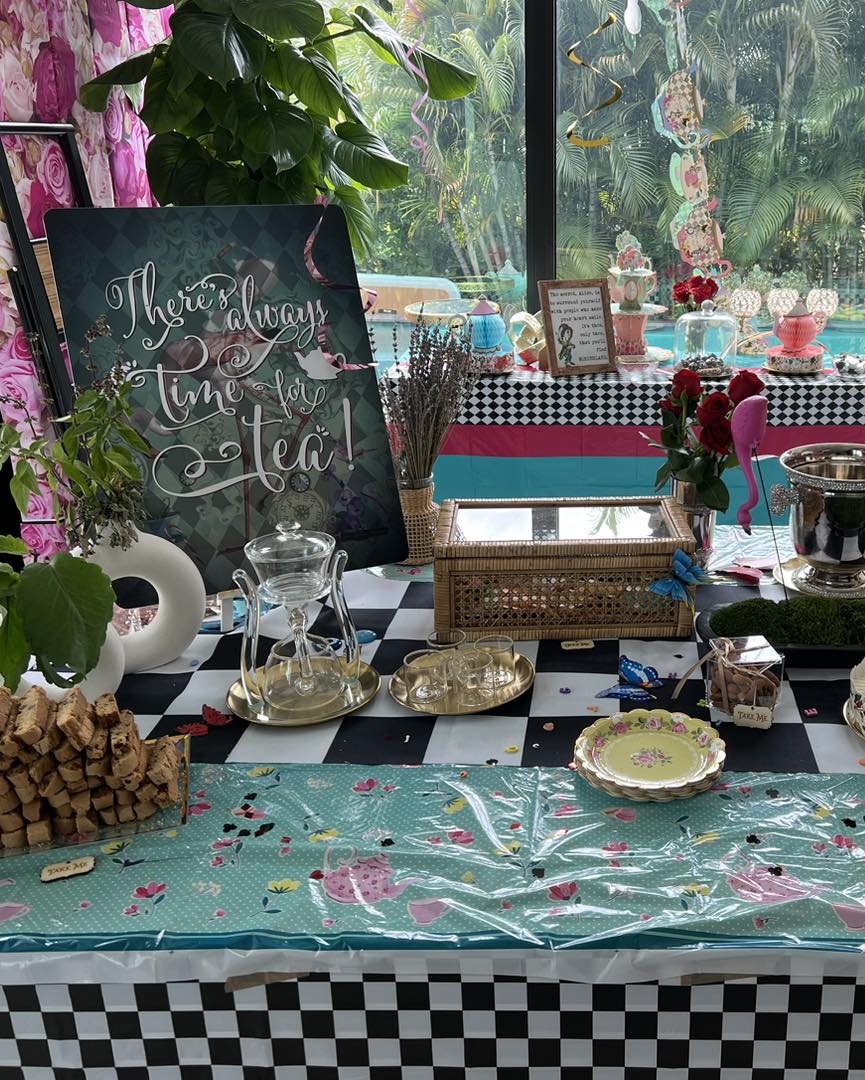 Elegant Alice in Wonderland party setup with a "there's always time for tea" sign, decorated tables with pastries and teapots, and a tropical backdrop.