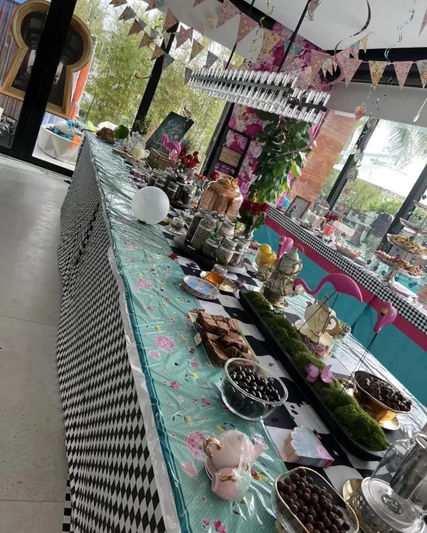 An indoor party table decorated with a whimsical tea party theme, featuring a checkered tablecloth, assorted sweets, and tea set accessories.