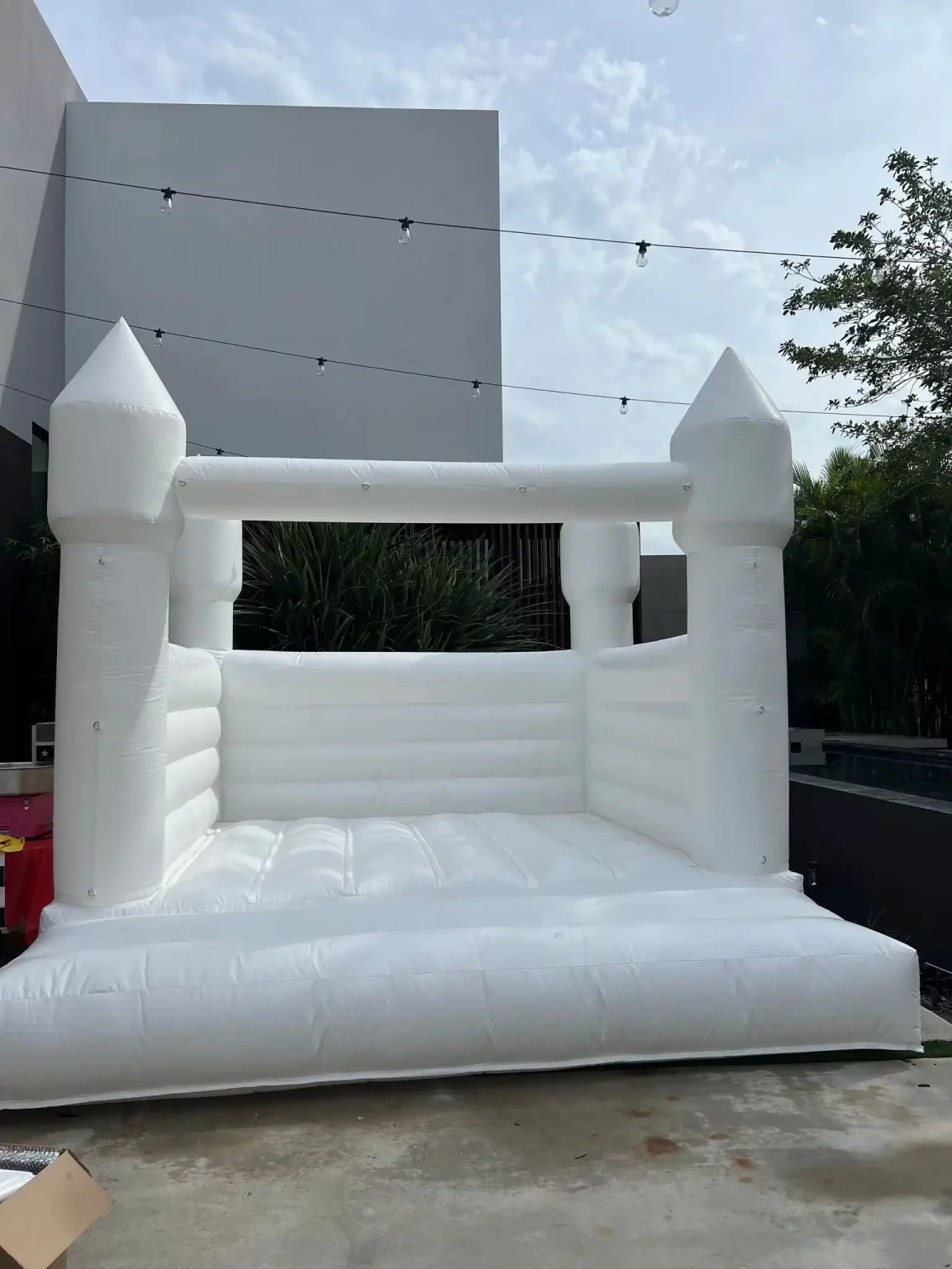 Inflatable white castle-shaped bouncy house with no roof, set up on a concrete surface near a building with string lights above.