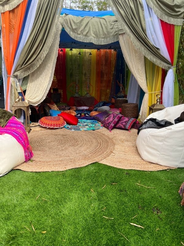 Colorful outdoor tent with vibrant curtains and an array of cushions and bean bags on a circular woven mat, set on a grassy surface for an Alice in Wonderland party.