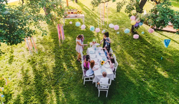 Aerial view of a children's outdoor birthday party. Adults and kids gather around a decorated table from a Kids Table and Chair Rental in a sunny garden.