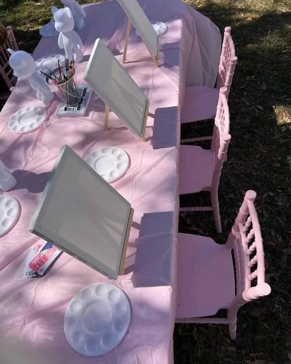 Outdoor Painting Party with pink chairs, canvases on easels, and palettes on a table, set up on grass under sunlight.