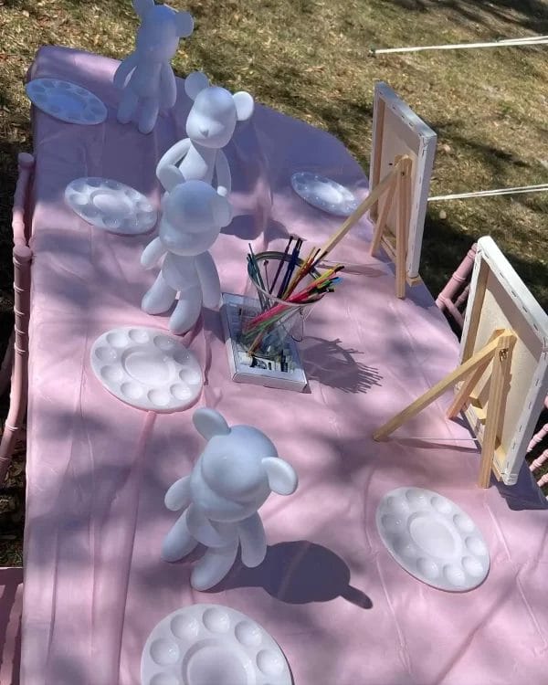 Outdoor Painting Party station with blank vinyl figures, palettes, and painting supplies on a pink tablecloth.
