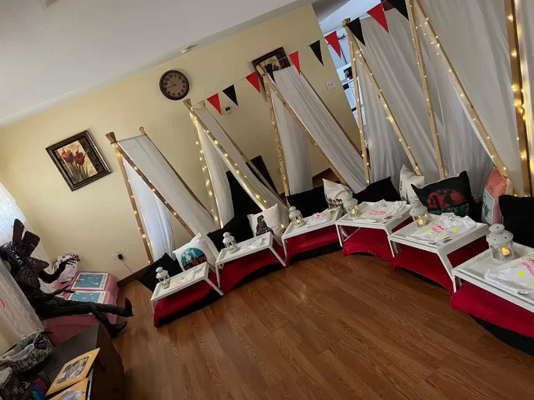 Indoor teepee sleepover setup with white draped tents, fairy lights, and Wednesday Addams Party-themed bedding on red floor mats in a living room.