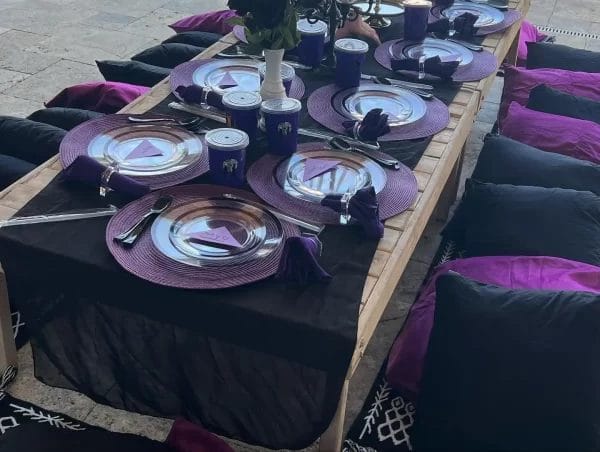Outdoor dining table set for six with purple placemats and cushions, featuring plates, cutlery, and glasses on a wooden table, perfect for a Wednesday Addams Party.
