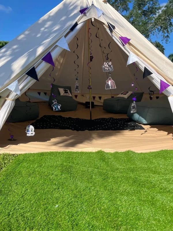 A large Wednesday Addams Party tent with open flaps, adorned with purple triangle flags and Addams Family decorations, set up on a grassy area under clear skies.