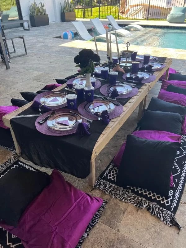 Outdoor dining setup with a rustic wooden table, purple plates and cushions, set near a pool with a slide and a dog in the background, designed for an Wednesday Addams Party.