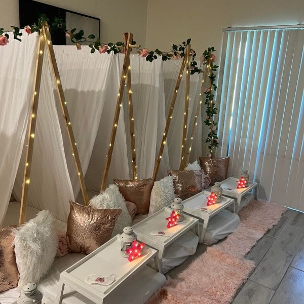 Indoor picnic setup with glam-themed party tents adorned with fairy lights and roses, floor cushions, and trays with candles, against a backdrop of vertical blinds.