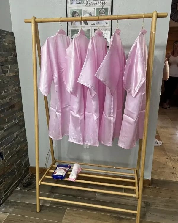 A wooden rack with five pink robes hanging in a room with a smiling woman partially visible at a Spa Party with DIY Lip Gloss in the background.