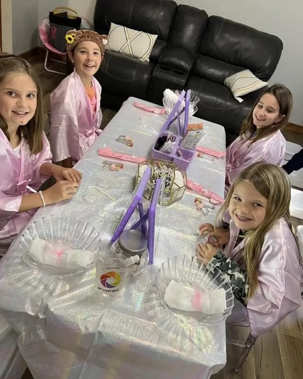 Four girls in robes sitting at a table, smiling, ready for a Spa Party with DIY Lip Gloss items scattered around.