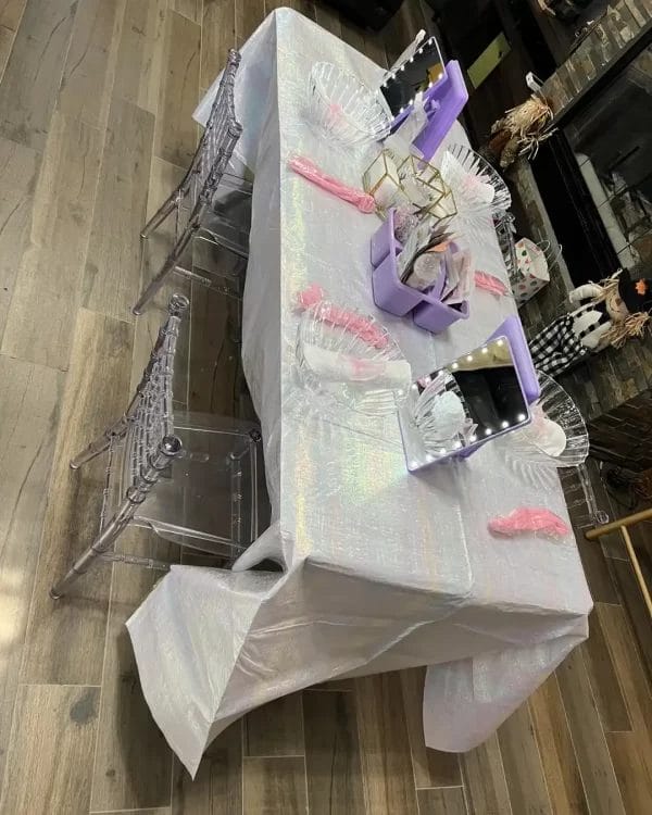 A cluttered display table covered with iridescent film, featuring a variety of items including a purple binder, clear chairs, and assorted glassware for a Spa Party with DIY Lip Gloss.