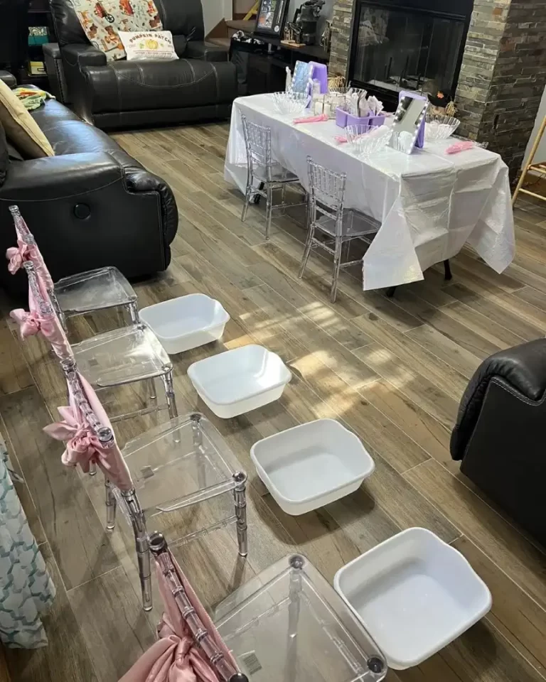 Transparent chairs with pink accents arranged around tables with white cloths and purple decorations in a cozy room with a stone fireplace and wooden flooring, prepared for a Spa Party with DIY lip Gloss event.