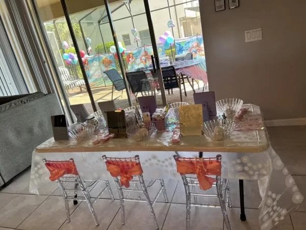 A children's birthday party setup indoors with a themed Spa Party with DIY lip Gloss tablecloth, transparent chairs with orange bows, and a pool visible through glass doors. The theme of the party is a Spa Party with DIY Lip Gloss Party.