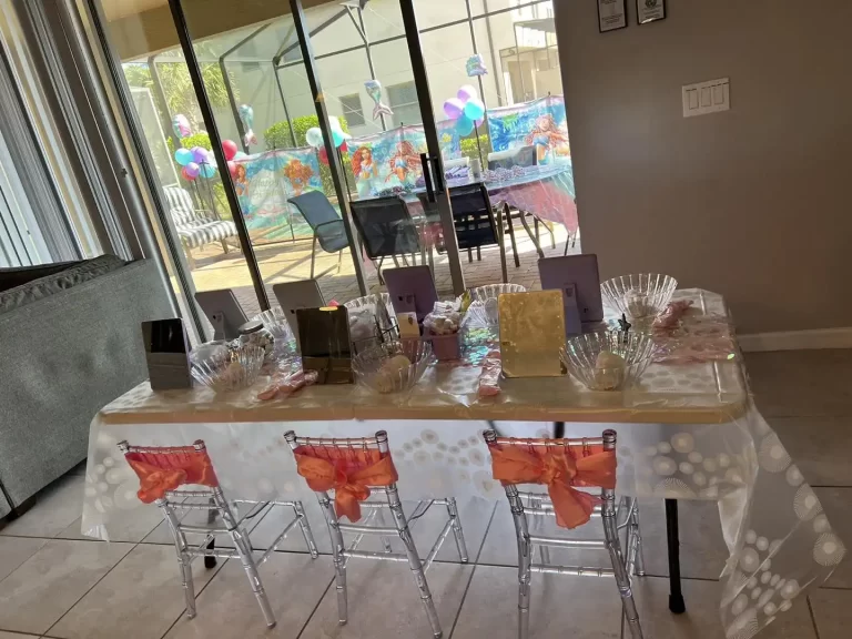A children's birthday party setup indoors with a themed Spa Party with DIY lip Gloss tablecloth, transparent chairs with orange bows, and a pool visible through glass doors. The theme of the party is a Spa Party with DIY Lip Gloss Party.