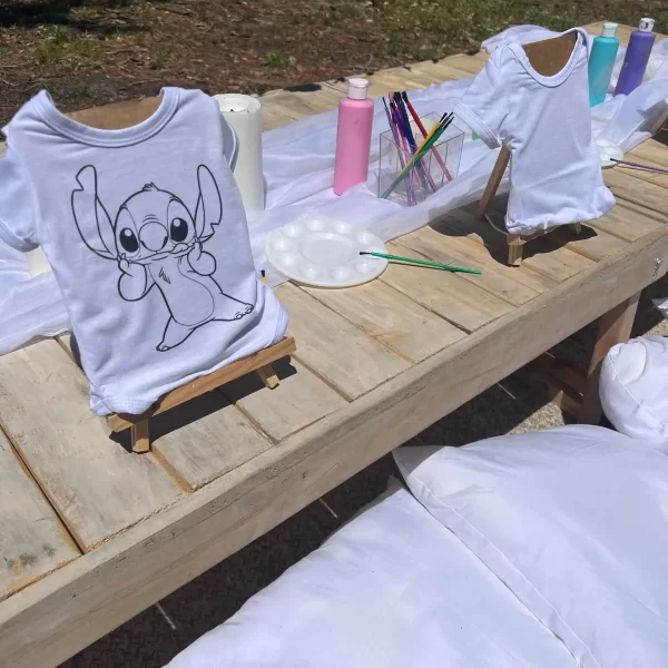 A white t-shirt with a sketched design of Painting Party displayed on a wooden stand at an outdoor art party with fabric paints.