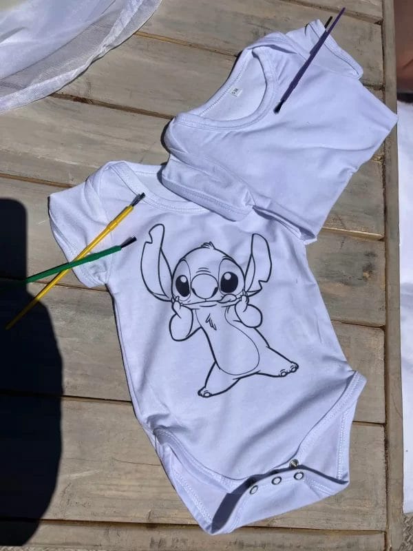 A white baby onesie with a printed design of Stitch from "Lilo & Stitch" laying on a wooden surface with two colored pencils, ideal for a Painting Party.