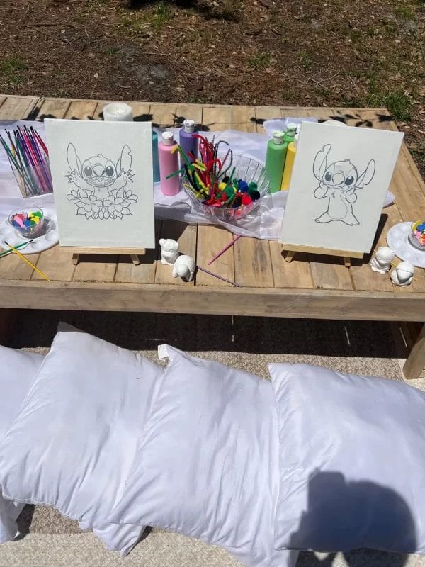 Outdoor Painting Party table with supplies, including brushes, colors, and two sheets of paper with animal drawings, located next to a bench with white cushions.
