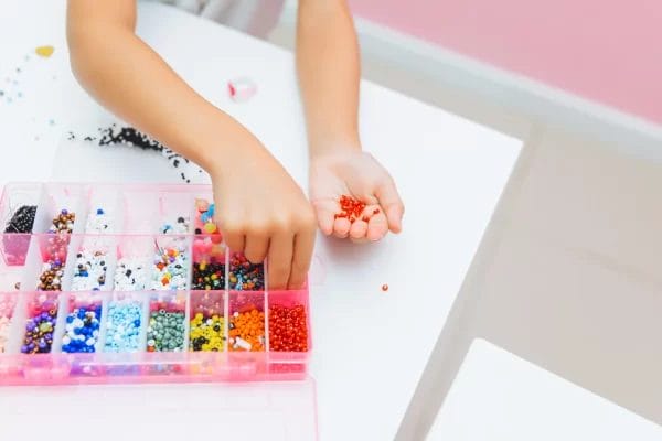 A child's hands selecting red beads from a colorful assortment for a Jewelry Art Party project in a storage box on a white table.