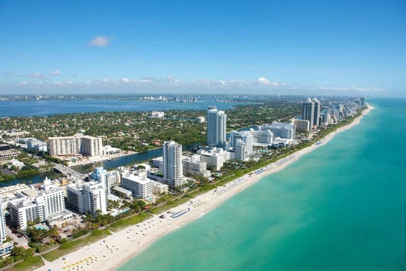 Aerial view of a coastal city with towering skyscrapers along a turquoise beach, extending towards a clear horizon.