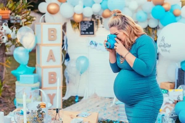 Pregnant woman in a blue dress smiling while taking a photo at a baby-themed party with decorations and gifts during a Crafty Onesie Painting Party for Your Baby Shower.