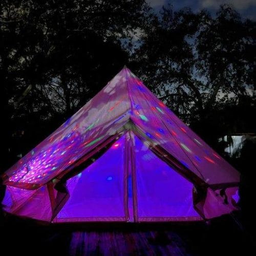 A Bell Tent lit up at night with purple lights, creating a magical and vibrant party atmosphere for glamping enthusiasts. The tent's fabric reflects colorful light patterns, accentuating its triangular shape.