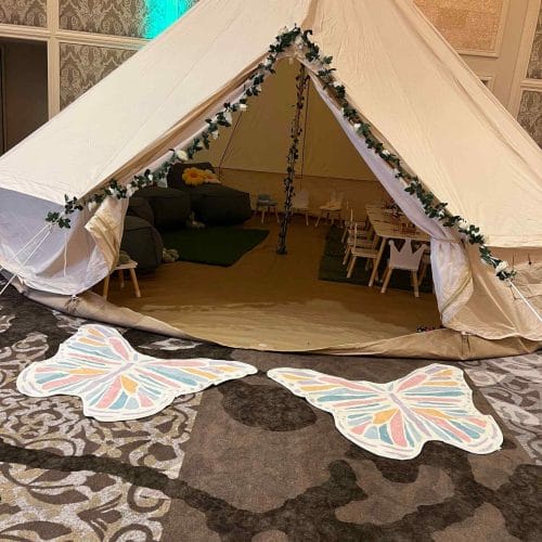 A large indoor tent decorated with leafy garlands at the entrance, flanked by two butterfly-shaped mats on a patterned carpet, perfect for various party themes.